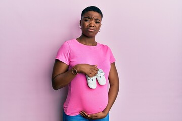 Poster - Young african american woman expecting a baby holding shoes clueless and confused expression. doubt concept.