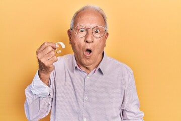 Senior man with grey hair holding medical hearing aid scared and amazed with open mouth for surprise, disbelief face