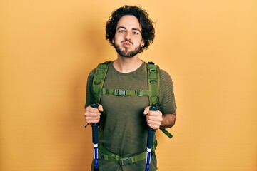 Canvas Print - Handsome hispanic man wearing backpack holding trekking poles puffing cheeks with funny face. mouth inflated with air, catching air.