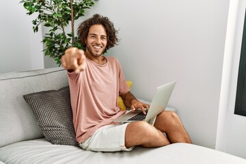 Canvas Print - Young hispanic man sitting on the sofa at home using laptop pointing to you and the camera with fingers, smiling positive and cheerful