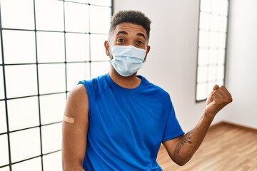 Wall Mural - Young hispanic man with beard wearing safety mask getting vaccine screaming proud, celebrating victory and success very excited with raised arms