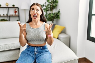 Sticker - Young brunette woman sitting on the sofa at home crazy and mad shouting and yelling with aggressive expression and arms raised. frustration concept.