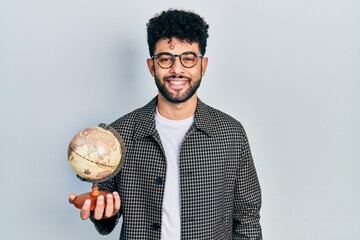 Sticker - Young arab man with beard holding vintage world ball looking positive and happy standing and smiling with a confident smile showing teeth