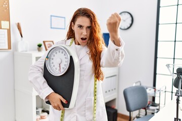 Poster - Young redhead woman nutritionist doctor holding weighing machine annoyed and frustrated shouting with anger, yelling crazy with anger and hand raised