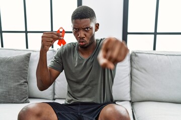 Poster - Young african american man holding support red ribbon sitting on the sofa pointing with finger to the camera and to you, confident gesture looking serious