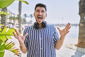 Poster - Young handsome man listening to music using headphones outdoors crazy and mad shouting and yelling with aggressive expression and arms raised. frustration concept.