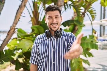 Poster - Young handsome man listening to music using headphones outdoors smiling friendly offering handshake as greeting and welcoming. successful business.