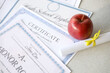 A honor roll recognition, certificate of achievement and high school diploma lies on table with small scroll and red apple. Education documents