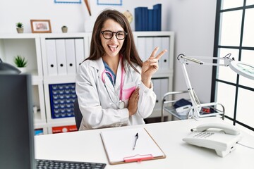 Wall Mural - Young doctor woman wearing doctor uniform and stethoscope at the clinic smiling with happy face winking at the camera doing victory sign. number two.