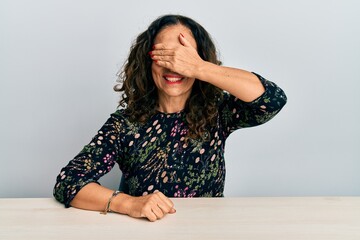 Canvas Print - Beautiful middle age woman wearing casual clothes sitting on the table smiling and laughing with hand on face covering eyes for surprise. blind concept.