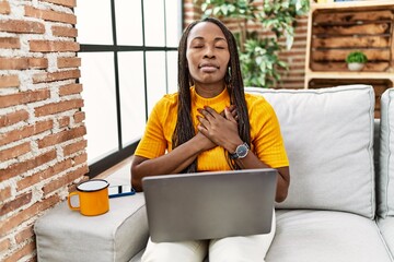 Wall Mural - African woman sitting on the sofa using laptop at home smiling with hands on chest with closed eyes and grateful gesture on face. health concept.