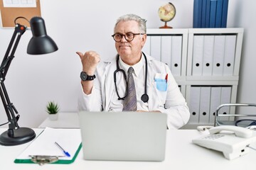 Wall Mural - Senior caucasian man wearing doctor uniform and stethoscope at the clinic smiling with happy face looking and pointing to the side with thumb up.