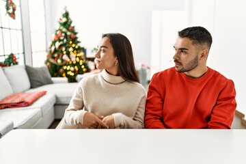 Canvas Print - Young hispanic couple sitting on the table by christmas tree looking to side, relax profile pose with natural face with confident smile.