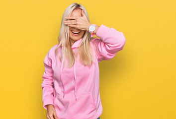 Poster - Young blonde woman wearing casual sweatshirt smiling and laughing with hand on face covering eyes for surprise. blind concept.