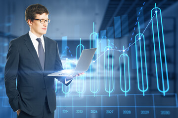 Poster - Attractive young european businessman using laptop with growing annual business chart report hologram on blurry office interior background. Analysis and company concept. Double exposure.