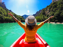 Back View Of A Woman With Hat Outstretching Arms For Happiness Sitting On A Red Canoe Kayak In A Blue River Tour. Outdoor Tourist Leisure Activity On Travel Vacation