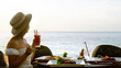 © TravelMedia - Young woman have romantic breakfast in sunrise at resort fine dining restaurant outdoor enjoy vacation looking at sea. Beautiful girl wear hat hold fruit cocktail. Healthy food drink for breakfast.