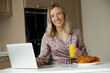 © Sergey - A young smiling girl in a home dress eats croissants and drinks orange juice and works at a laptop