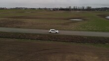 White Car 2019 Toyota Corolla Hatchback Driving Along Country Farm Road Surrounded By Grass Brown Fields Mountains In Farmland Abbotsford BC Aerial Wide Leading Tracking Car Accelerates Side Profile