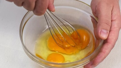 Wall Mural - Male hands beat eggs in a glass bowl a whisk on a white table. Close up.