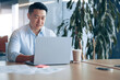 © Yaroslav Astakhov - Smiling Asian businessman working on laptop at her workplace in modern office. Business concept