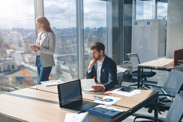 businessman analyzes work papers and charts while his colleague looks thoughtfully out the window. 