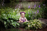Fototapeta  - Malutki chłopiec w czapeczce misia siedzi w ogrodzie pomiędzy fioletowymi kwiatami i pięknie się uśmiecha