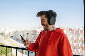 Wall Mural - Young happy handsome man wearing red hoodie doing video call using smartphone with headphone. Man watching video on phone or video call or scrolling on social media.
