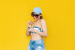 A young caucasian smiling blonde woman in a swimsuit top, blue cap, denim shorts, sunglasses trying sorbet, holding a cream bowl with ice cream isolated on a color yellow background. Summer concept