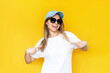 © Марина Демешко - A young caucasian cheerful smiling blonde woman in a blue cap and sunglasses pointing at white casual t-shirt with fingers showing empty copy space for design and text on a color yellow background