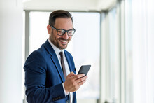 Handsome Businessman Wearing Eyeglasses Using Smart Phone And Smiling While Standing Near The Office Window, Copy Space