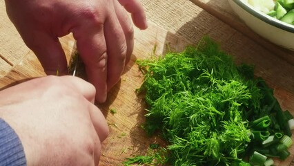 Wall Mural - a hand with a knife cuts dill on a wooden table for salad, close-up