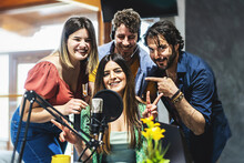 A Team Of Four Presenters Greets The Audience Of Their Live Streaming Vlog Podcast Via The Internet - A Group Of Content Creator Influencers Speak Into The Microphone During A Live Broadcast - 