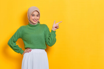 Poster - Cheerful beautiful Asian woman in green sweater pointing fingers aside at copy space isolated over yellow background