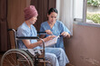 © tigercat_lpg - Uniformed young Asian female therapy doctor motivates wheelchair male patient at window by reading book to support recovery cancer illness after chemo medical treatment in hospital inpatient room.