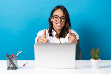 Wall Mural - Hispanic pretty young woman making ok sign in front of laptop isolated on blue background. Office worker on video conference