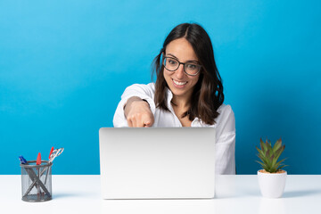 Wall Mural - Hispanic young woman pointing at webcam of laptop while sitting behind desk isolated on blue background.