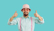© Studio Romantic - Cheerful funny charismatic young man showing thumbs up isolated on turquoise background. Bearded hipster in shirt, suspenders, panama and sunglasses looking at camera with funny expression on his face