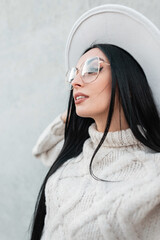 Wall Mural - Urban female portrait of beautiful woman model with white hat with vintage glasses in trendy knitted sweater on a gray wall on the street