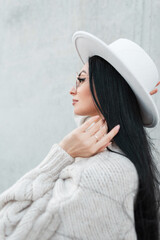 Wall Mural - Fashionable beautiful young woman with long brunette hair in a trendy white vintage sweater with a hat and glasses on a gray concrete background