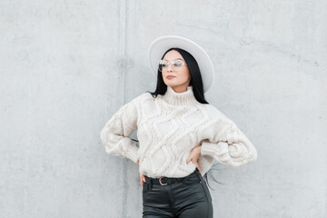 Wall Mural - Happy beautiful young girl with fancy vintage glasses and fashionable clothes with a sweater, hat, and leather pants stands near a concrete gray wall on the street