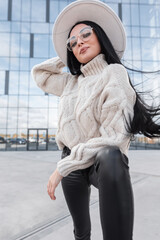 Wall Mural - Fashion trendy young Caucasian woman with long hair and glasses in knitted vintage sweater, white hat and leather pants poses in the city. Modern female urban style outfit