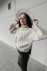 Wall Mural - Happy Caucasian woman with smile and vintage glasses in trendy beige knit sweater and hat walks in the city