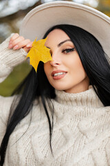 Wall Mural - Beauty autumn portrait of fashion happy young Caucasian woman model with smile and blue eyes in stylish knitted vintage sweater and hat covers her eye with a yellow fall leaf outdoors