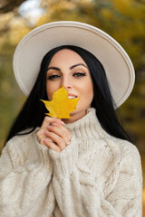 Wall Mural - Happiness pretty girl with smile in vintage fashion sweater and white round hat holds a yellow autumn leaf and walks in the fall park. Trendy woman looks at the camera