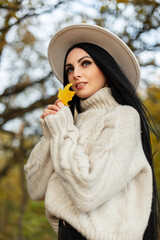 Wall Mural - Happy beautiful young woman in a fashionable vintage knit outfit with beige sweater and hat is holding an autumn yellow leaf and walking in the park