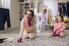 Busy Woman Has Rubber Pink Gloves On Hands. Mother Of Two Daughters Is Cleaning Bathroom On Knees Scrubbing Floor With Polish Brush And Talking On Phone. Little Girls Are Helping Their Mom Clean.