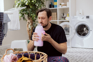 Wall Mural - Laundry room, bathroom. Man dressed in household clothes sits on floor by wicker basket with things to wash. Man holds laundry detergent in hands and sniffs its scent. A wry face, he doesn't like it.