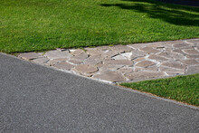 Asphalt Road Near Garden Path Made Of Natural Stone Paved With Rough Rock In A Park With A Green Lawn, The Decorative Path With An Abstract Pattern.