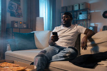 A man's studio in the evening illuminated by street light coming through the window, cigarette smoke, man with dark skin, relaxed, sitting on the couch watching TV, holding a phone in hand